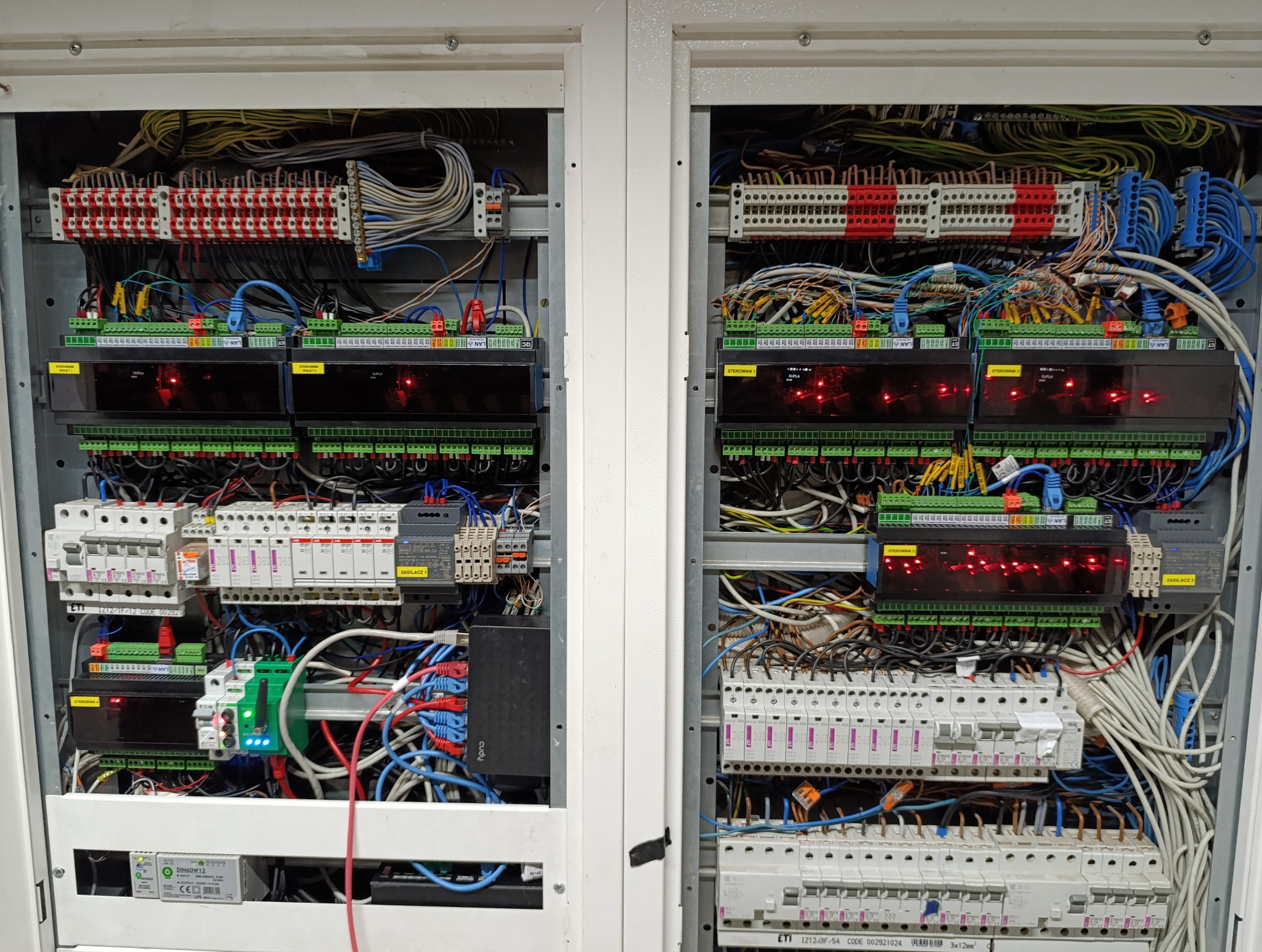 Dwie otwarte szafy sterownicze z widocznymi modułami sterującymi, bezpiecznikami, listwami zaciskowymi i okablowaniem w kolorach niebieskim, czerwonym, zielonym i żółtym. Czerwone diody LED świecą...