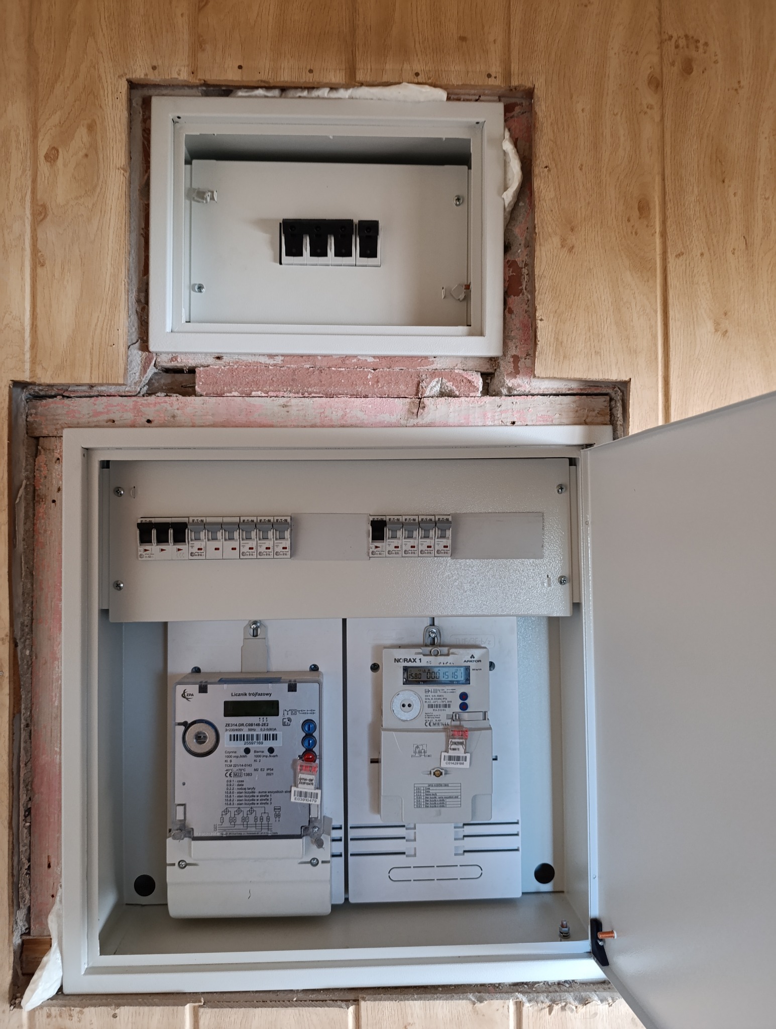 Otwarta skrzynka elektryczna z dwoma licznikami trójfazowymi, bezpiecznikami i pianką montażową uszczelniającą obudowę w drewnianej ścianie.