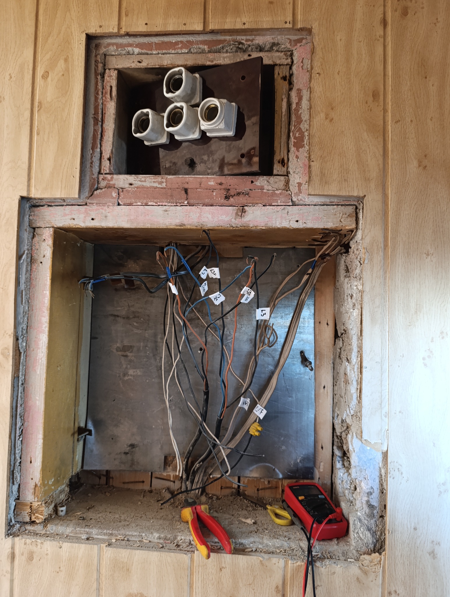 Otwarta skrzynka elektryczna w ścianie z widocznymi przewodami oznaczonymi etykietami, ceramicznymi gniazdami bezpieczników, multimetrem i obcęgami leżącymi na dnie skrzynki.