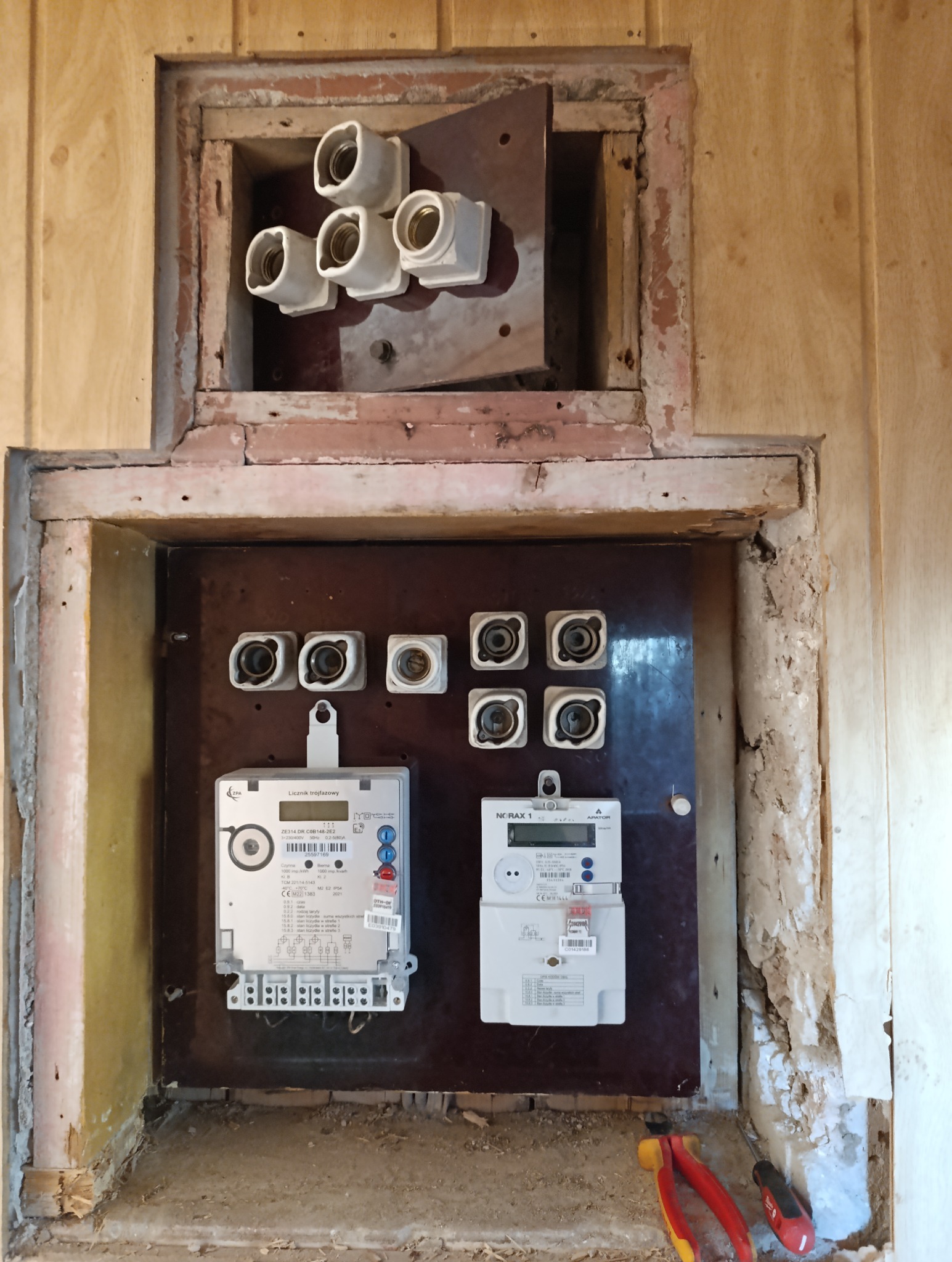 Skrzynka z dwoma licznikami elektrycznymi na brązowej płycie, powyżej bezpieczniki topikowe, ściany obudowy w trakcie remontu, widoczne narzędzia elektryczne.