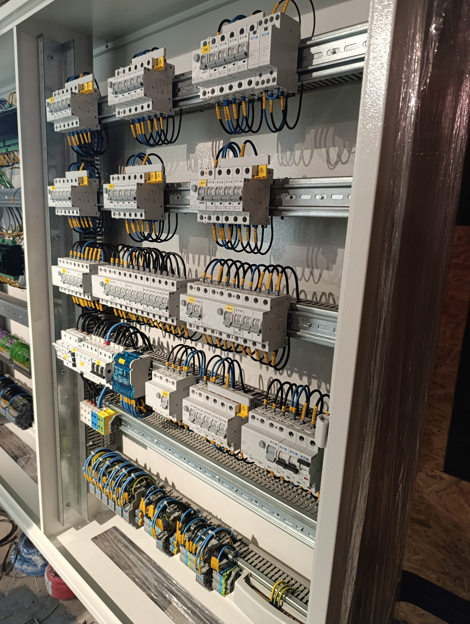 Rozdzielnia elektryczna z zamontowanymi bezpiecznikami Eaton, widoczne przewody w kolorach niebieskim i żółtym z oznaczeniami, szyny DIN oraz elementy montażowe w metalowej obudowie, częściowo...