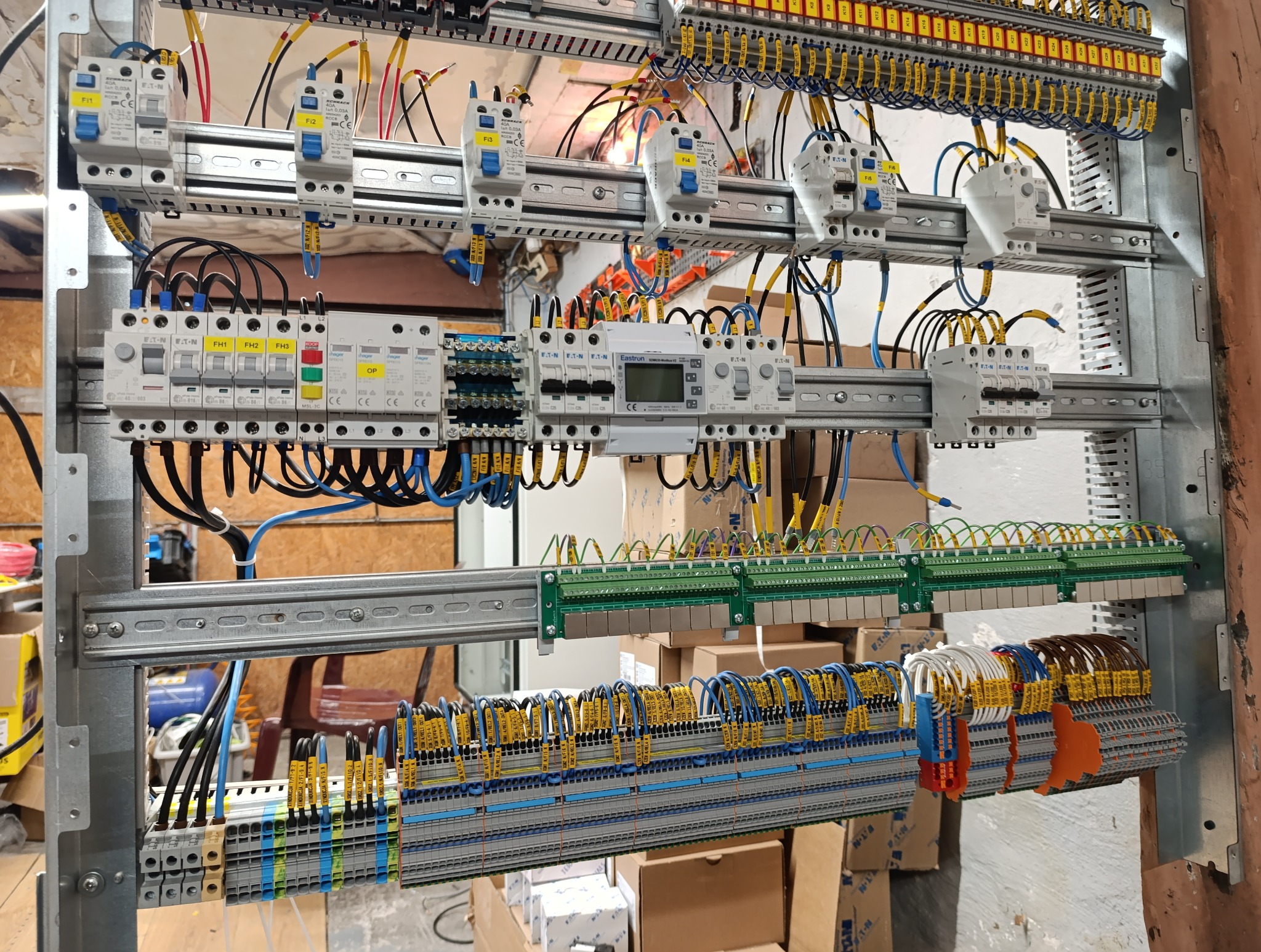 Rozdzielnia elektryczna z licznymi bezpiecznikami, przewodami oznaczonymi żółtymi etykietami i listwami zaciskowymi, zamontowana na metalowej konstrukcji w pomieszczeniu technicznym.