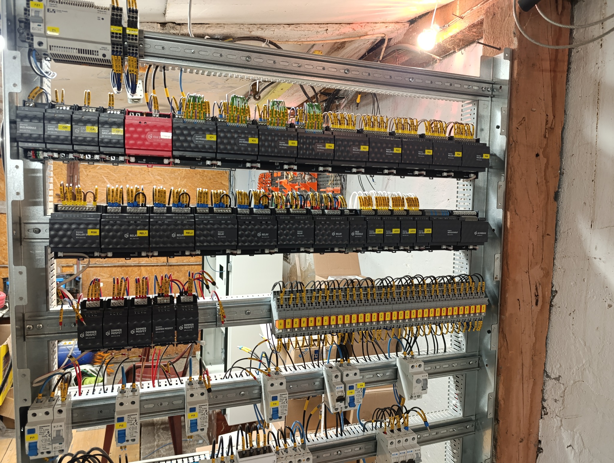 Rozbudowana szafa sterownicza z modułami Greton i Eaton, okablowana przewodami oznaczonymi żółtymi etykietami, zamontowana na metalowej konstrukcji przy ścianie z widocznym drewnianym elementem.