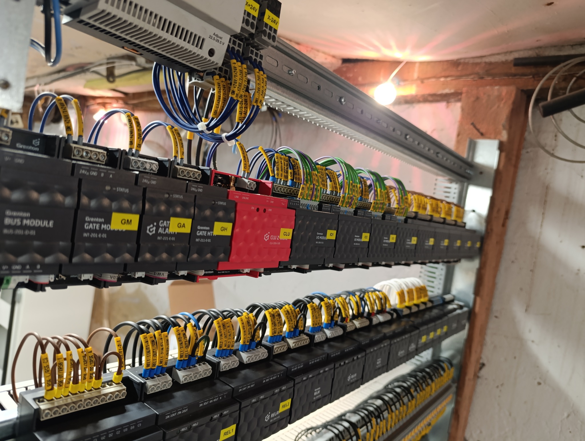 Rozbudowana szafa sterownicza z modułami Grenton, okablowana przewodami z żółtymi oznaczeniami, widoczne zasilacze, oświetlenie sufitowe w tle.