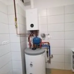 Nowoczesna kotłownia gazowa z białym piecem wiszącym i zasobnikiem ciepłej wody, rury miedziane prowadzone wzdłuż ściany z białych płytek, widoczne elementy armatury i zabezpieczeń.