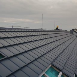Dekielski Dekarsko Ciesielski - Montaż ciemnoszarej blachodachówki modułowej na nowym dachu z widoczną więźbą i izolacją, pracownik w kasku na tle pochmurnego nieba.
