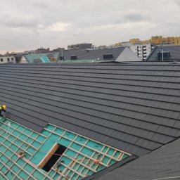 Dekielski Dekarsko Ciesielski - Pracownik w żółtej kamizelce układa ciemnoszarą dachówkę na nowej konstrukcji dachu z widoczną membraną izolacyjną i elementami więźby, z panoramą miasta w tle.