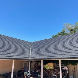 Dekielski Dekarsko Ciesielski - Nowo pokryty, ciemnoszary dach z dachówki ceramicznej, widoczny z perspektywy poddasza z pracującym mężczyzną na drabinie w tle, przy jasnym, błękitnym niebie.