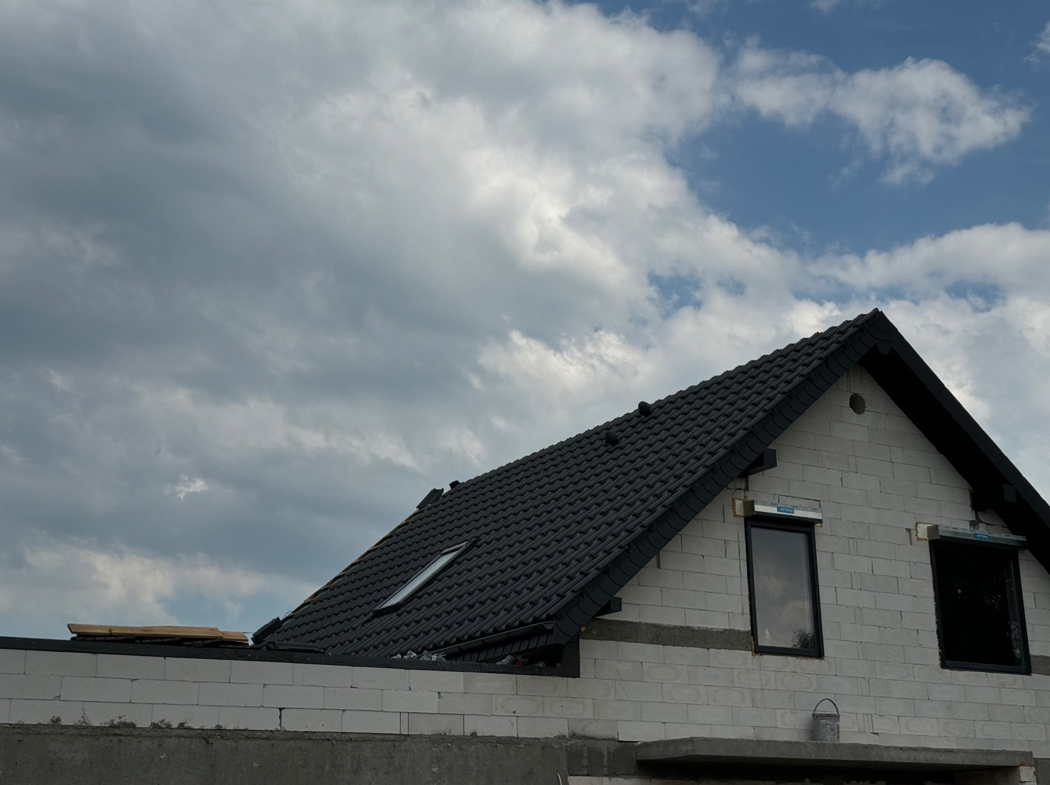 Dom w trakcie budowy z czarną dachówką i oknami z roletami zewnętrznymi, widok na dach i fragment ściany z betonu komórkowego na tle zachmurzonego nieba.