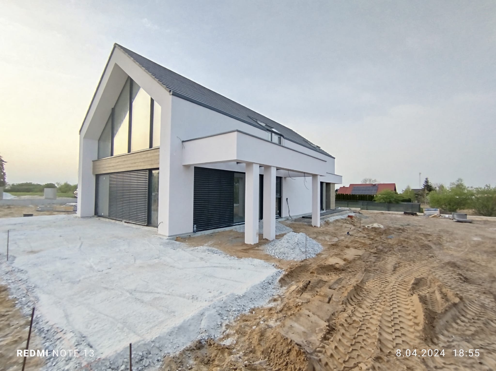 Nowoczesny dom jednorodzinny z grafitową dachówką, dużymi oknami i elementami wykończeniowymi w trakcie budowy, widoczne ślady opon i materiały budowlane.