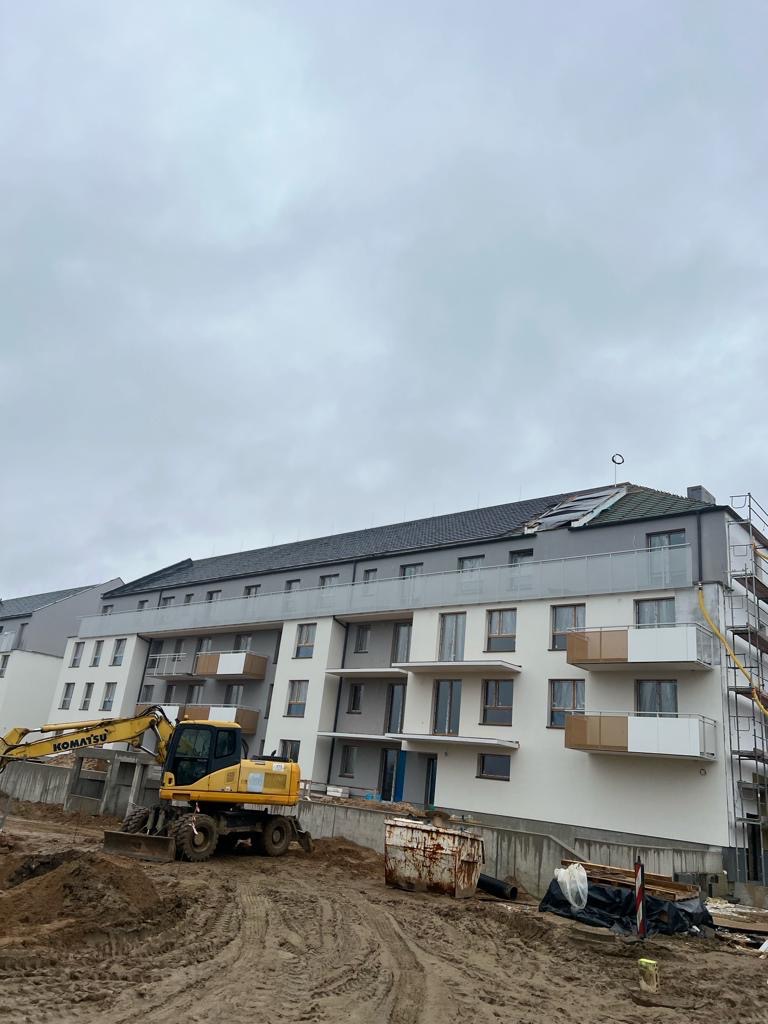 Nowy budynek wielorodzinny z szarą dachówką w trakcie budowy, widoczny żółty koparka Komatsu i rusztowania.