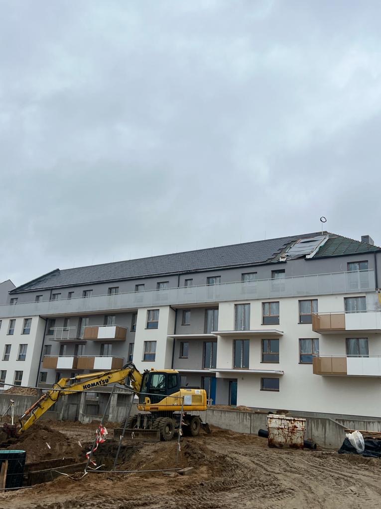Budynek wielorodzinny w trakcie budowy, widoczny fragment dachu z uszkodzeniami i żółta koparka Komatsu na pierwszym planie, teren budowy niezagospodarowany.