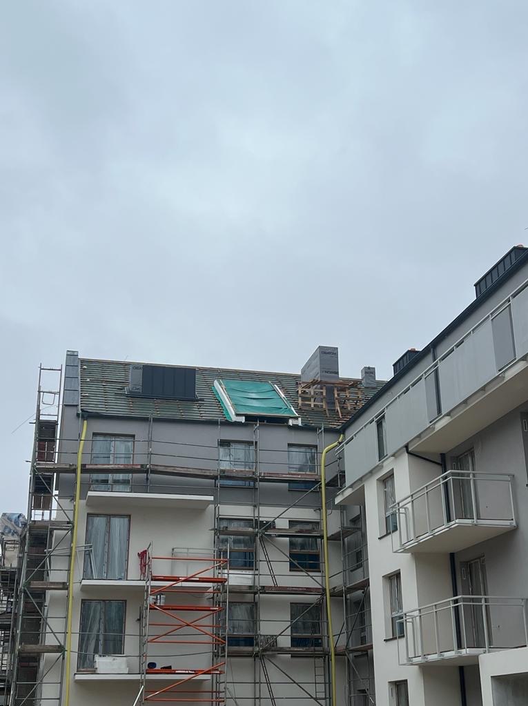 Wielopiętrowy budynek w trakcie remontu dachu, widoczne rusztowania, częściowo położona dachówka, fragment zabezpieczony plandeką, stos materiałów budowlanych na dachu.