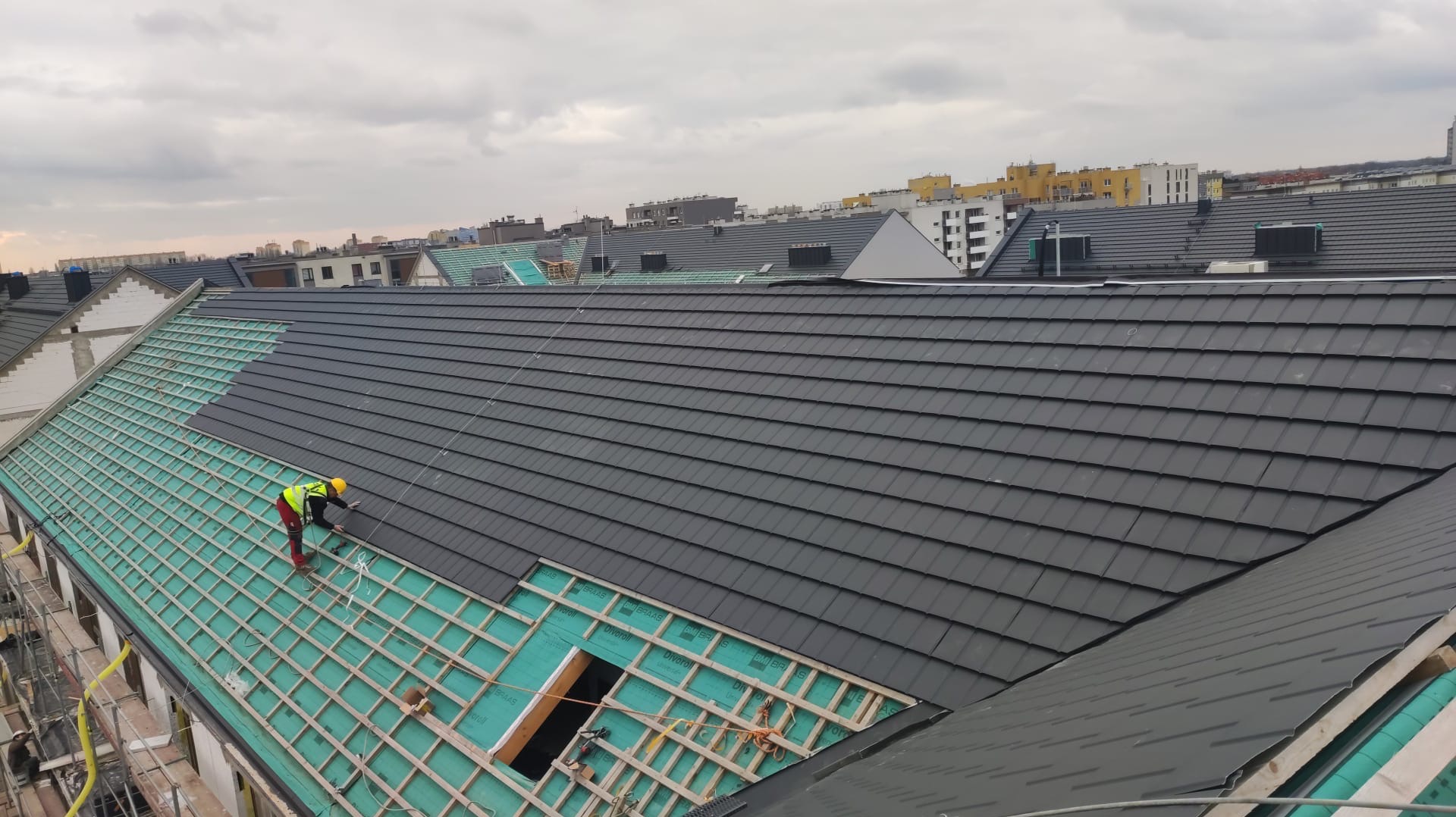 Pracownik w żółtej kamizelce układa ciemnoszarą dachówkę na nowej konstrukcji dachu z widoczną membraną izolacyjną i elementami więźby, z panoramą miasta w tle.