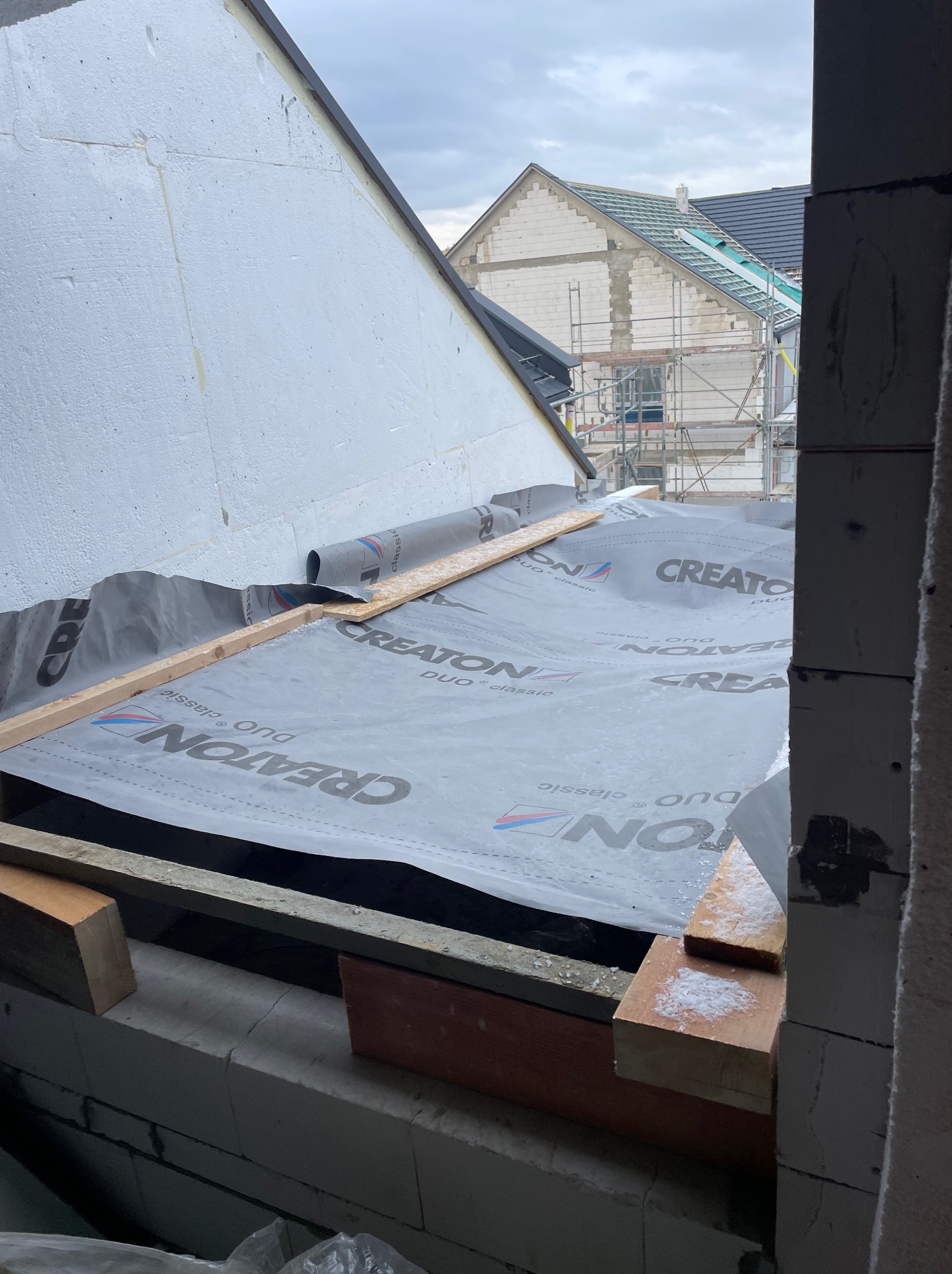 Widok z wnętrza budynku na membranę dachową marki Creaton Duo Classic, rozłożoną na drewnianej konstrukcji dachu w trakcie budowy, z widocznym ociepleniem i budynkiem w tle na rusztowaniu.