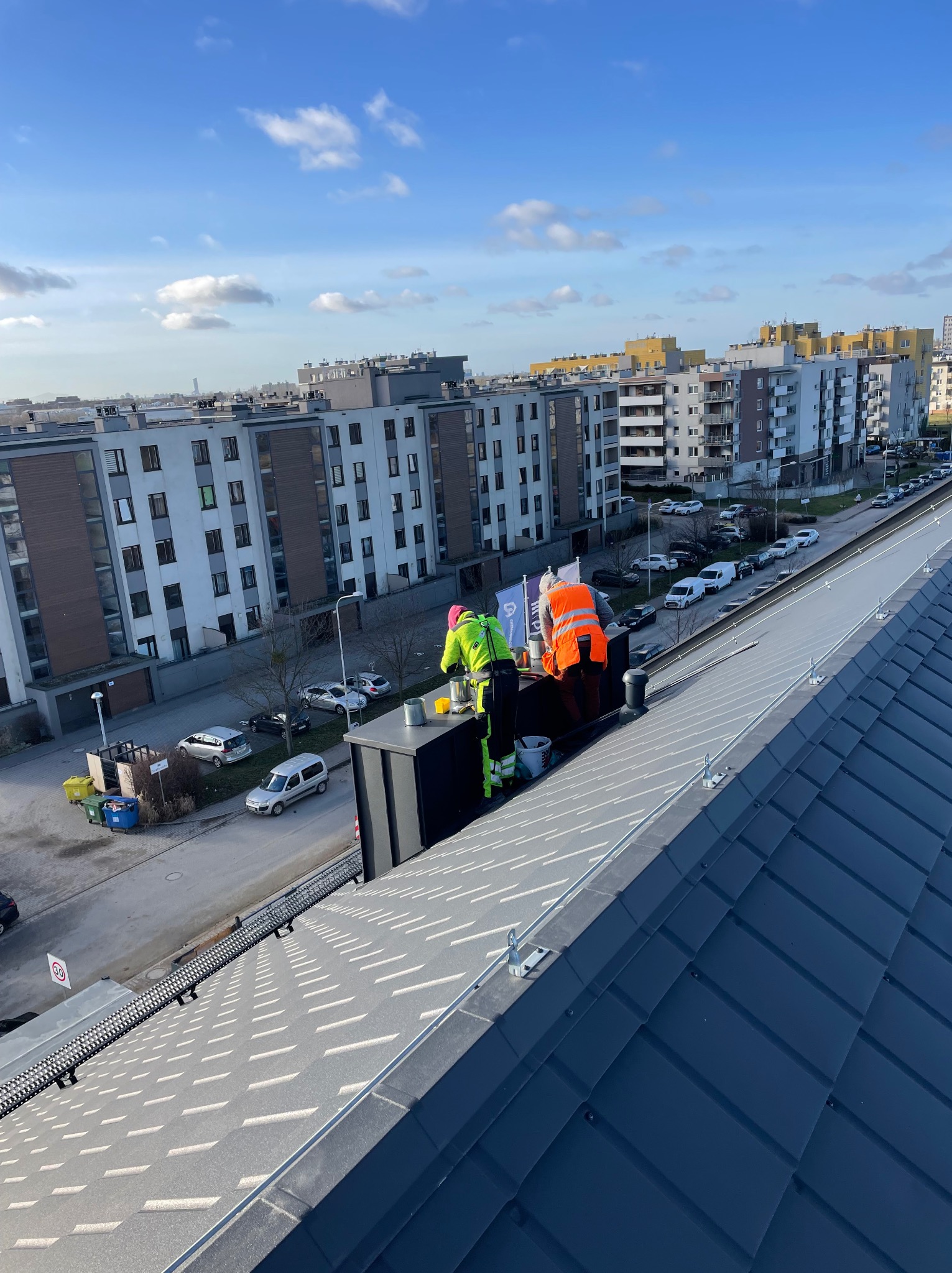 Dwóch pracowników w odblaskowych ubraniach na dachu budynku wielorodzinnego, montujących czarny komin wentylacyjny na tle zabudowy miejskiej w słoneczny dzień.