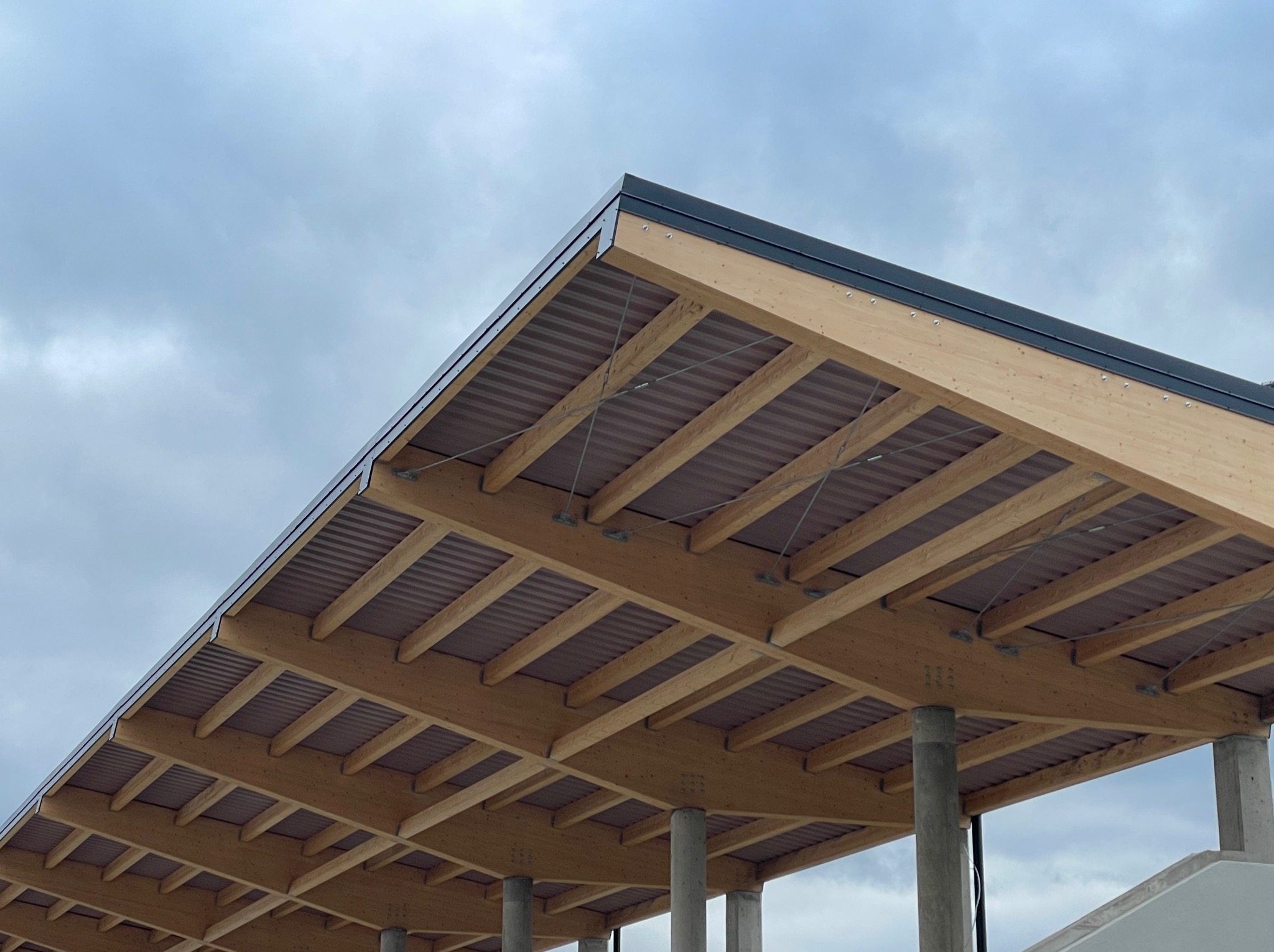 Drewniana konstrukcja zadaszenia z widocznymi elementami więźby, metalowym wykończeniem krawędzi i betonowymi słupami podtrzymującymi na tle pochmurnego nieba.