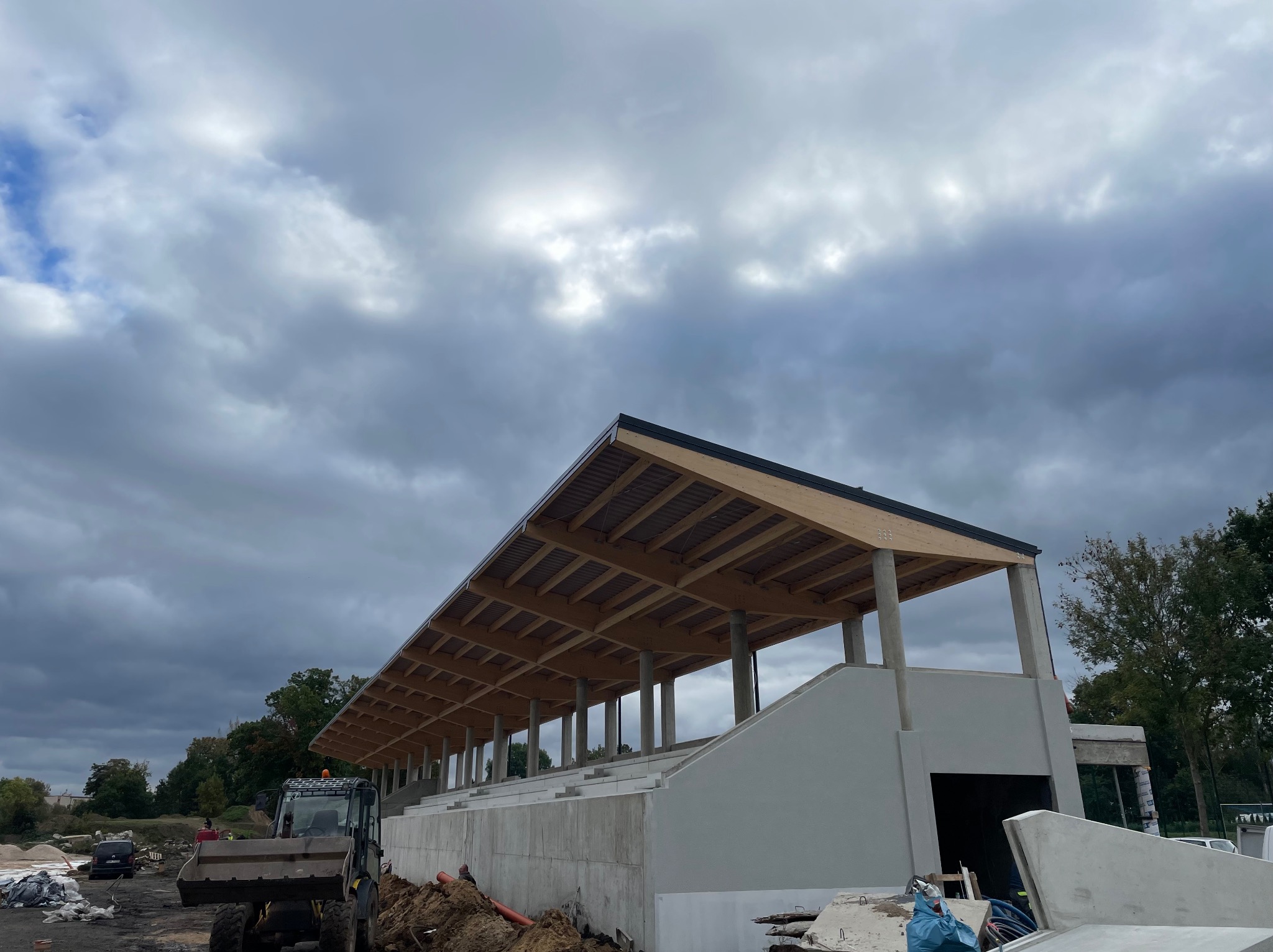 Budowa zadaszenia trybun sportowych z drewnianą konstrukcją dachu, widoczny traktor i plac budowy pod pochmurnym niebem.