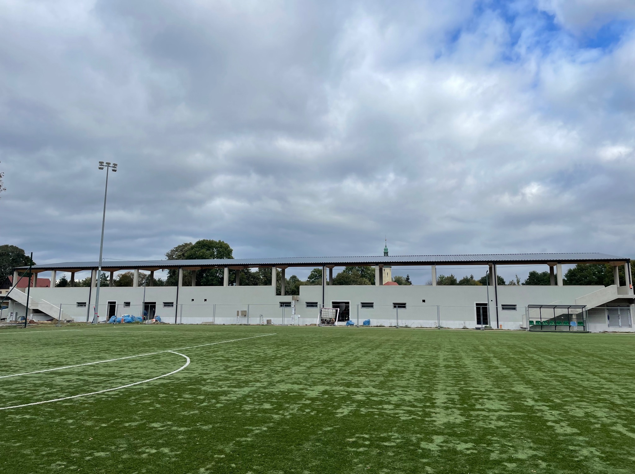 Nowo wybudowany budynek zadaszenia trybun na boisku sportowym z sztuczną trawą, widoczne kolumny podtrzymujące dach i szary metalowy dach, w tle wieża kościelna.