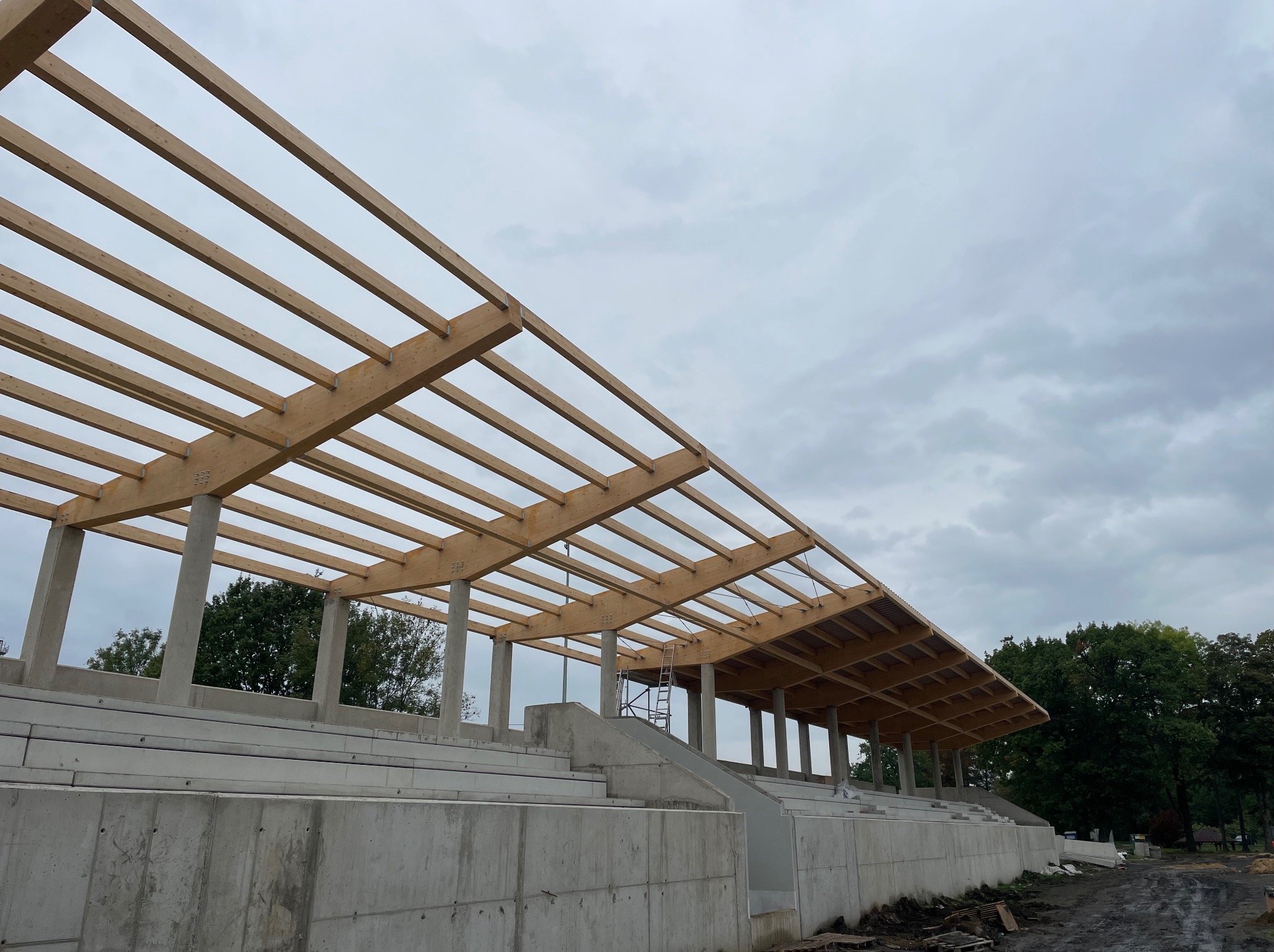 Konstrukcja z drewnianych belek tworząca zadaszenie trybun stadionu, wsparta na betonowych słupach i fundamentach, widok w pochmurny dzień.