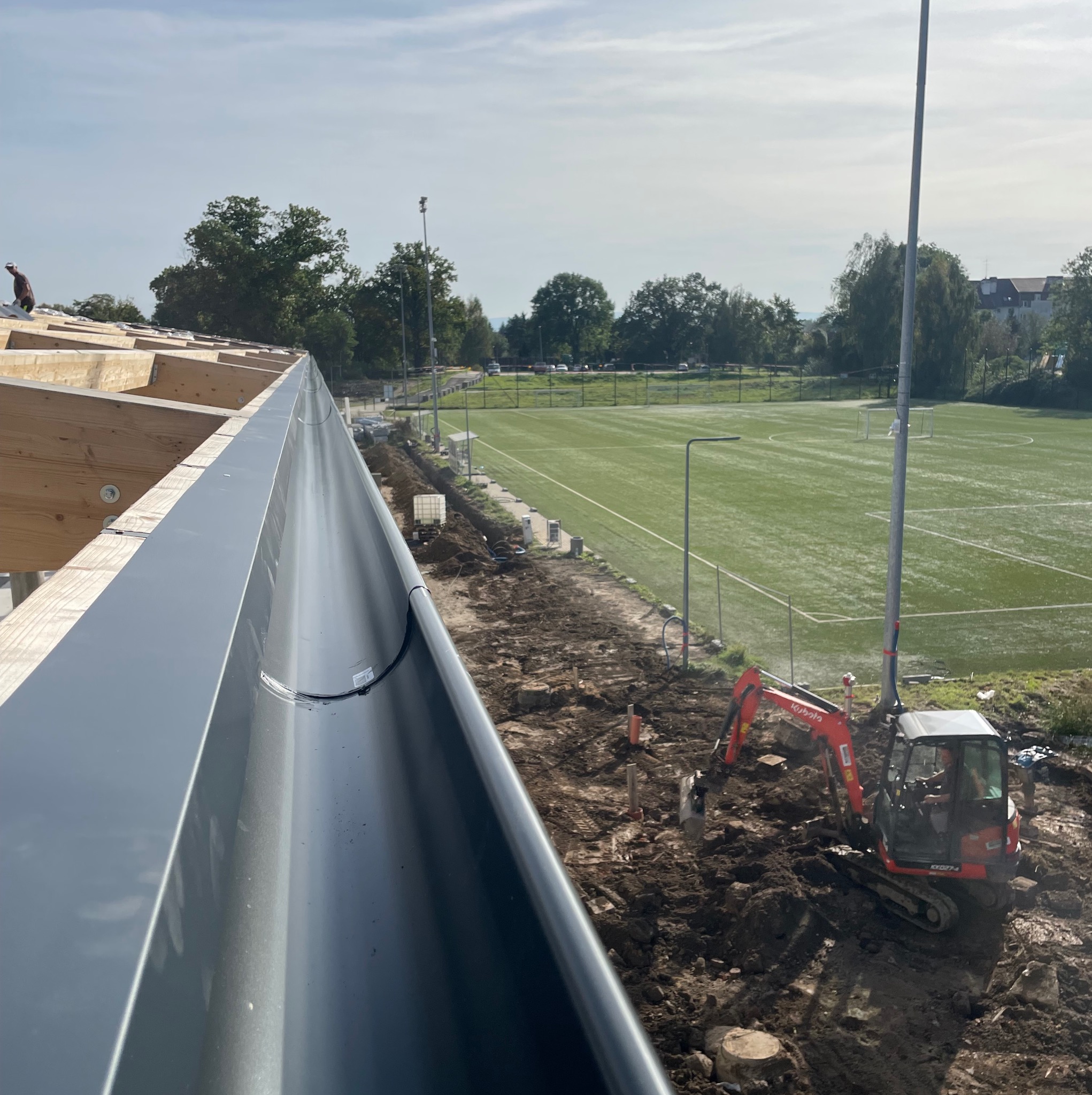 Widok z góry na zamontowaną, szarą rynnę z blachy na nowej więźbie dachowej z widocznym pracownikiem, w tle boisko sportowe i mała koparka Kubota na placu budowy.
