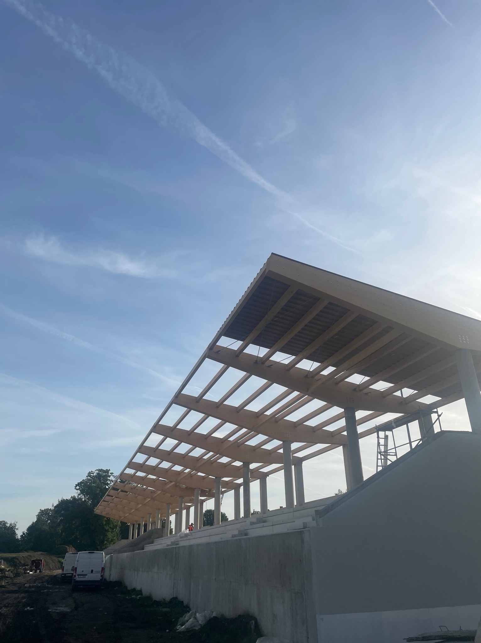 Konstrukcja z drewna klejonego na betonowym fundamencie, widoczne słupy i krokwie, błękitne niebo z smugami kondensacyjnymi w tle, budowa z bliska w słoneczny dzień.