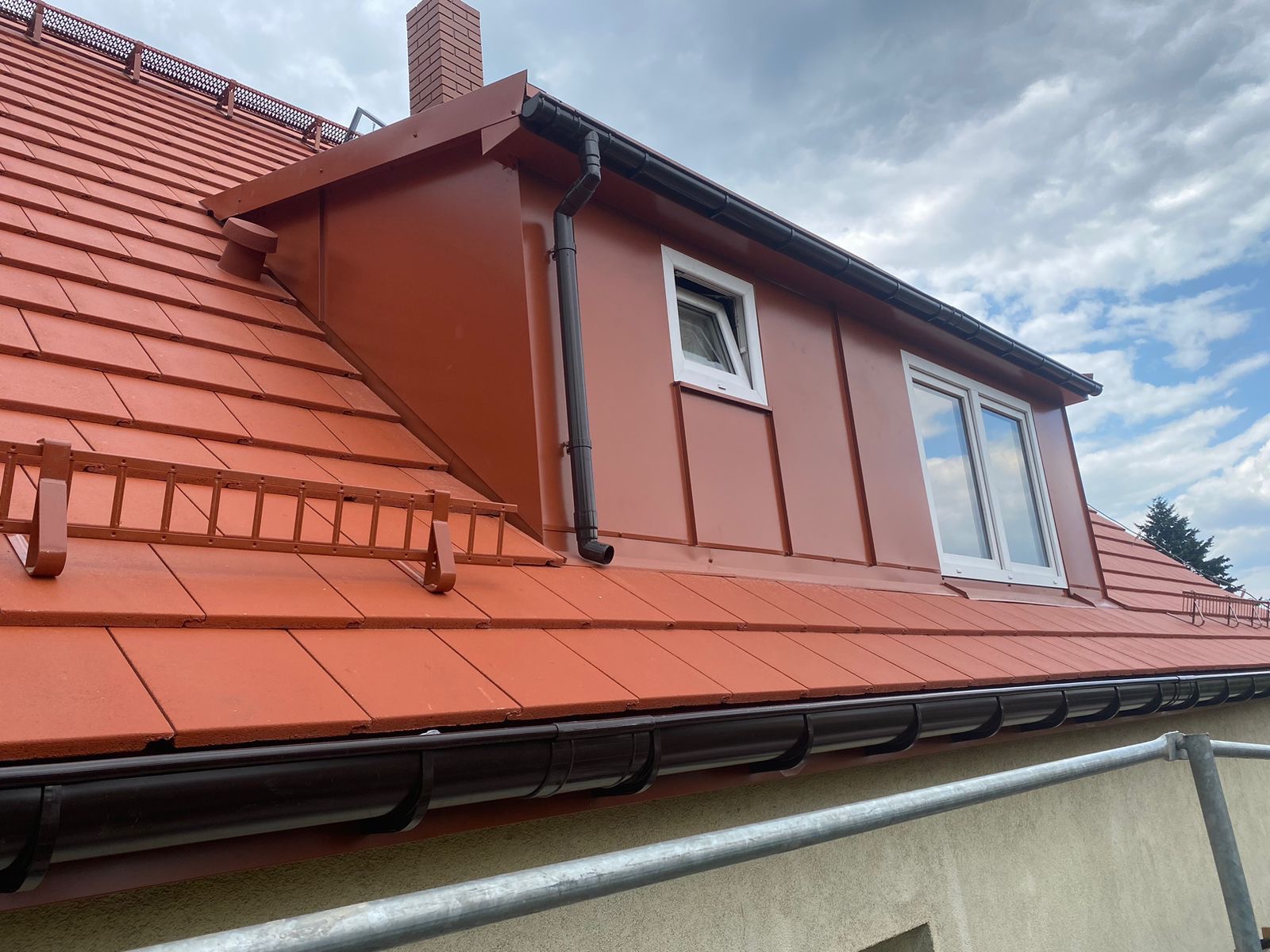Czerwony dach z dachówką ceramiczną, orynnowanie w kolorze grafitowym, okna dachowe z białymi ramami, komin z cegły, zabezpieczenie przeciwśniegowe w postaci drabinki.