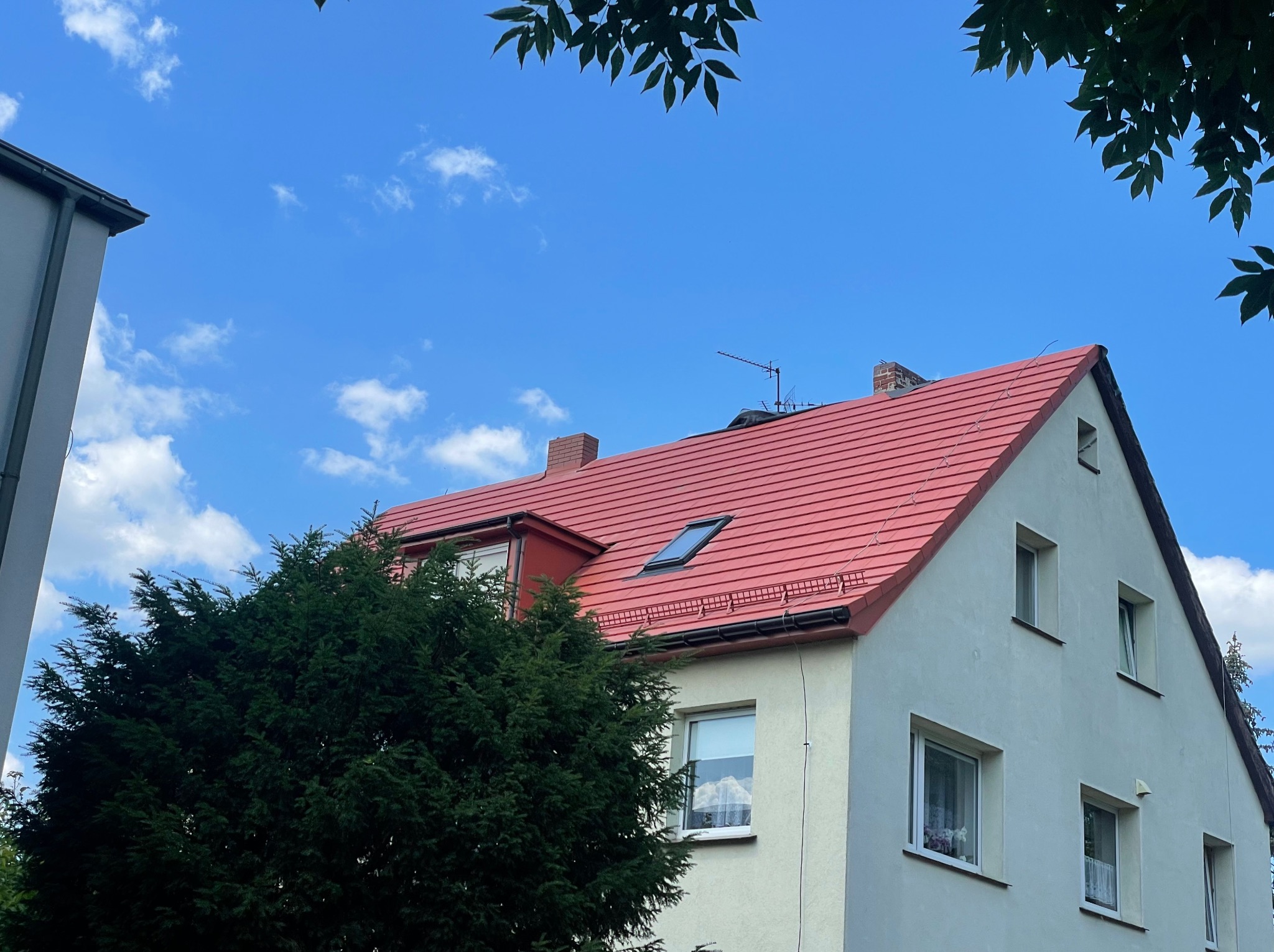 Dom jednorodzinny z nowym czerwonym dachem, widoczne okno dachowe i antena, częściowo zasłonięty przez iglaste drzewo.