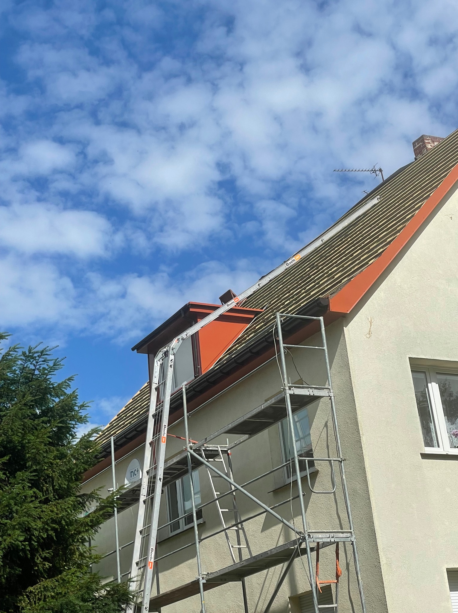 Dom jednorodzinny w trakcie prac dekarskich, widoczne rusztowanie, odkryta konstrukcja dachu z łatami, nowe orynnowanie w kolorze czarnym, błękitne niebo z chmurami w tle.