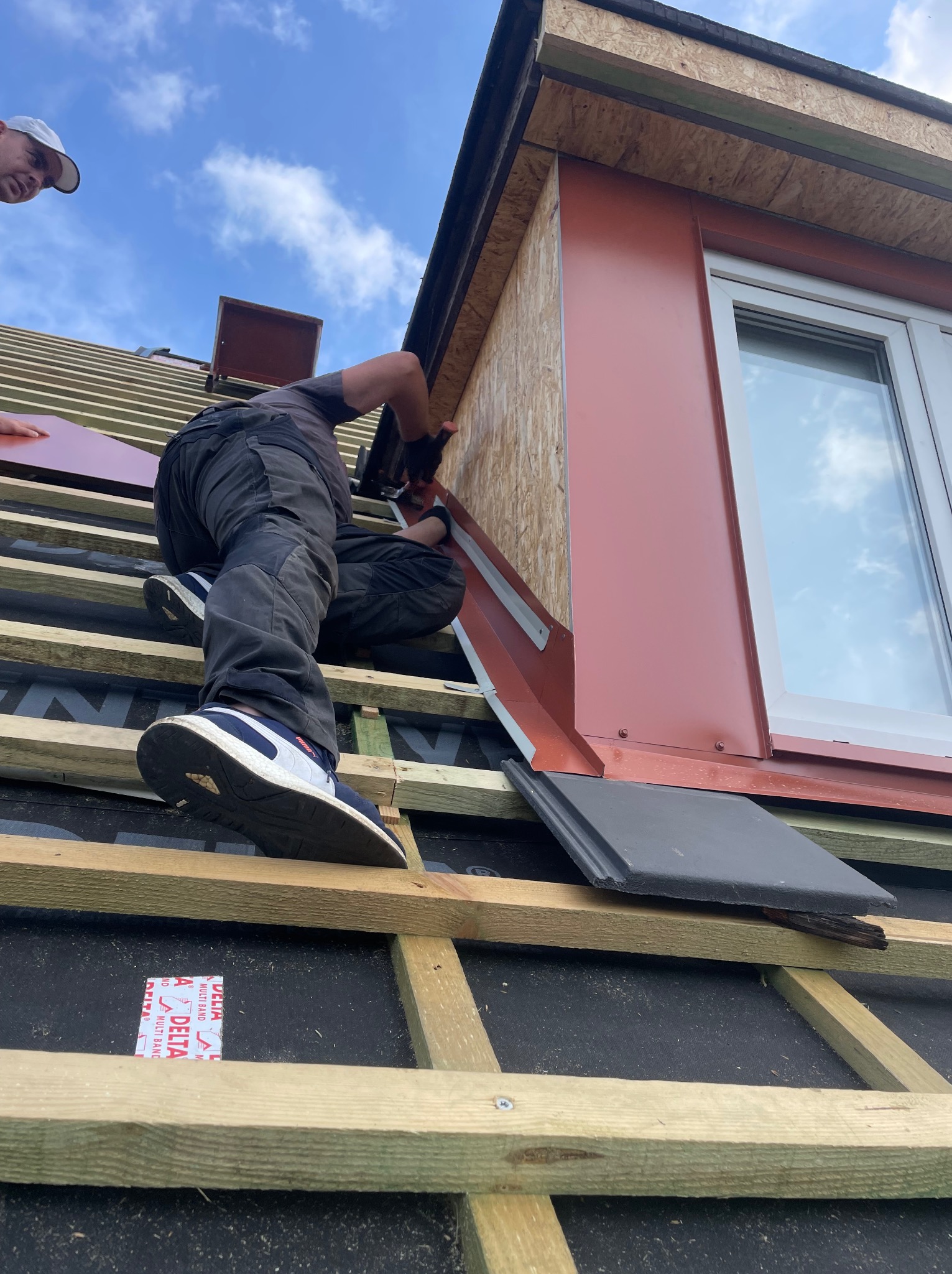 Monter w ciemnych spodniach i sportowych butach, z perspektywy od dołu, mocuje blaszane obróbki okna dachowego na drewnianej konstrukcji dachu, widoczna część nieba.