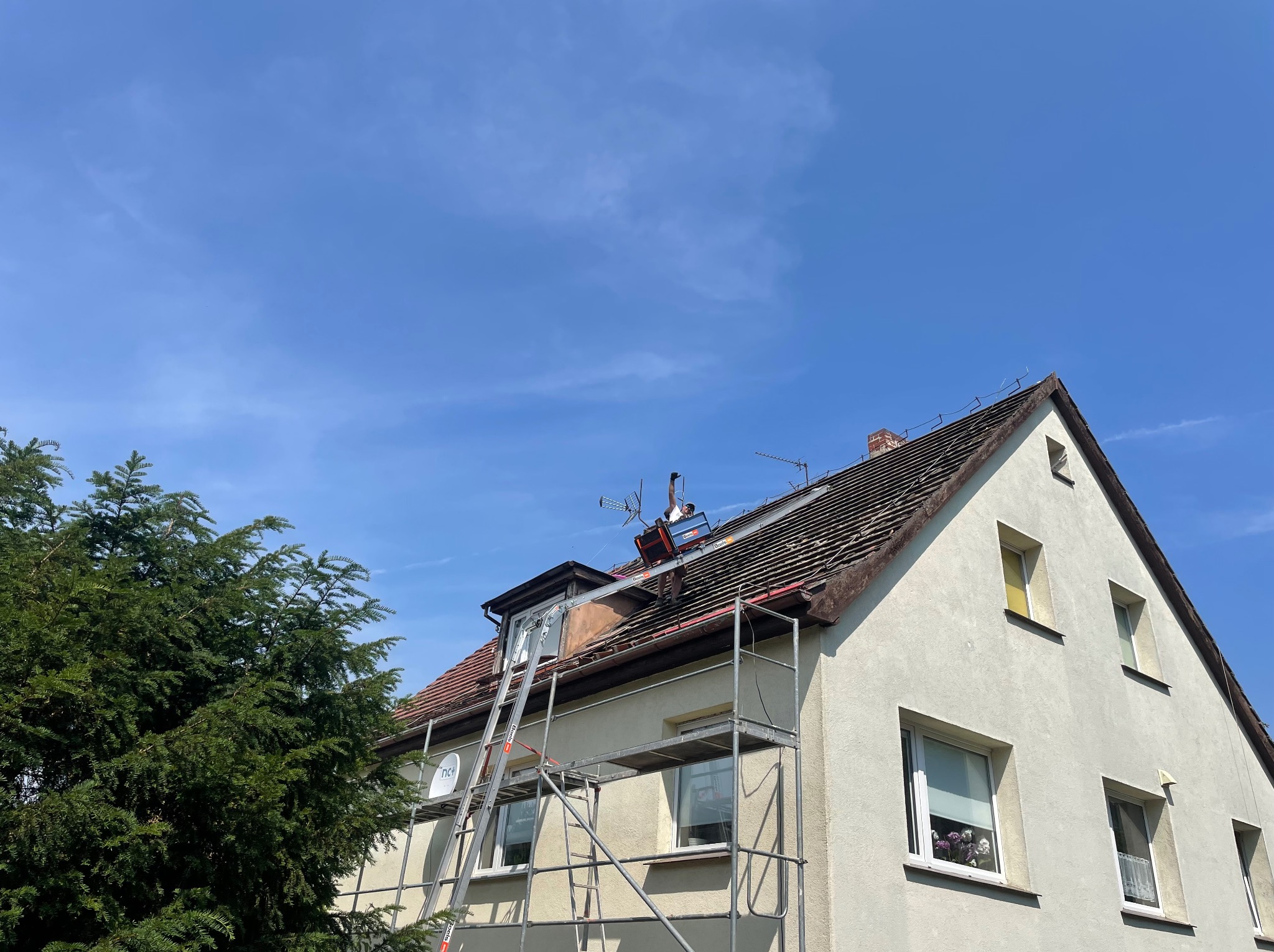 Pracownik na dachu domu jednorodzinnego zabezpieczony rusztowaniem, demontuje stare pokrycie w pobliżu okna dachowego, widoczne anteny satelitarne.