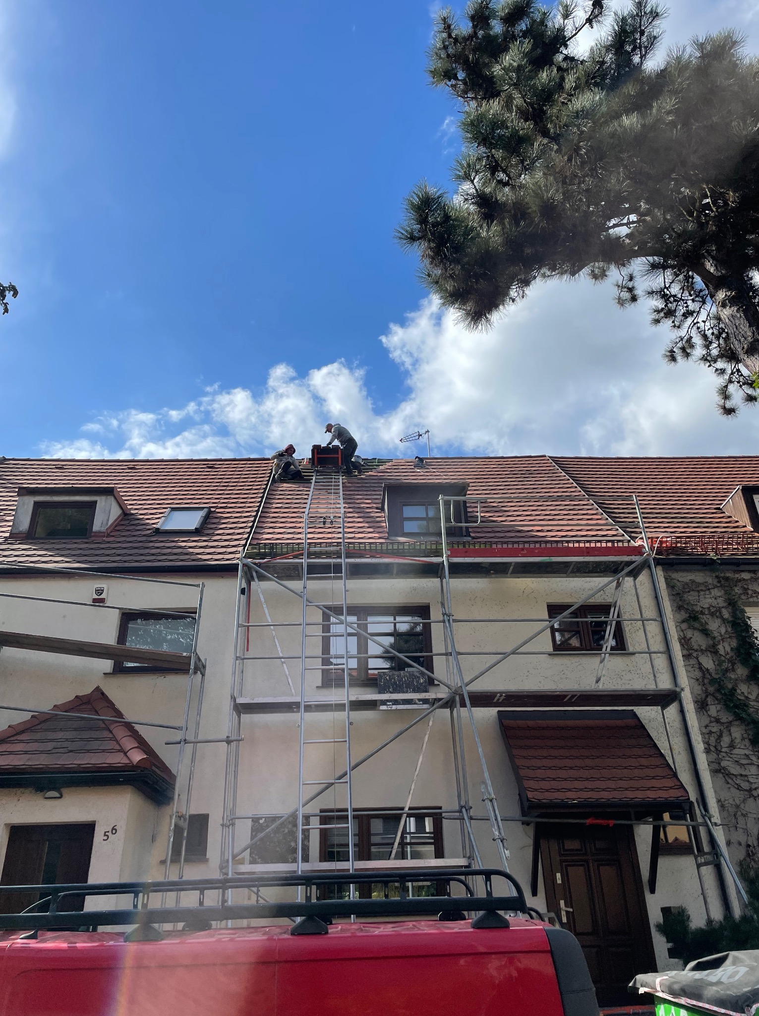 Dwóch dekarzy na rusztowaniu, pracujących na dachu dwupiętrowego domu z czerwonej dachówki, widok z dołu, fragment dachu samochodu w kadrze.