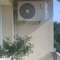 Biała jednostka zewnętrzna klimatyzacji Rotenso zamontowana na teksturowanej ścianie balkonu, widoczny krzew róży i fragment balustrady.