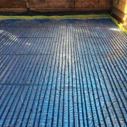 Niebieskie rury ogrzewania podłogowego zamocowane do czerwonej siatki na folii izolacyjnej, widok z góry w surowym wnętrzu.