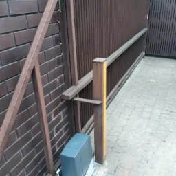 Brązowa brama przesuwna z zamontowanym szarym napędem automatycznym, widoczna listwa zębata i fragment muru z cegły.
