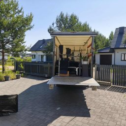 VELOX-TRANS KOMPLEKSOWE PRZEPROWADZKI - Otwarta paka busa wypełniona meblami i sprzętami podczas przeprowadzki. Na pierwszym planie zapakowany telewizor, w tle domy jednorodzinne i błękitne niebo.