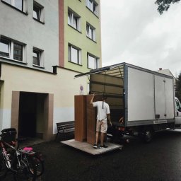 VELOX-TRANS KOMPLEKSOWE PRZEPROWADZKI - Mężczyzna w krótkich spodenkach wyładowuje owiniętą szafę z białej ciężarówki na tle bloku mieszkalnego. Obok zaparkowane rowery i ławka.