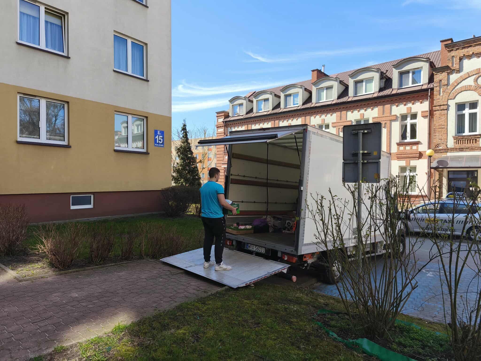 Mężczyzna w niebieskiej koszulce wnosi kartony do białego busa z otwartą rampą na tle budynku mieszkalnego z numerem 15. Słoneczny dzień.