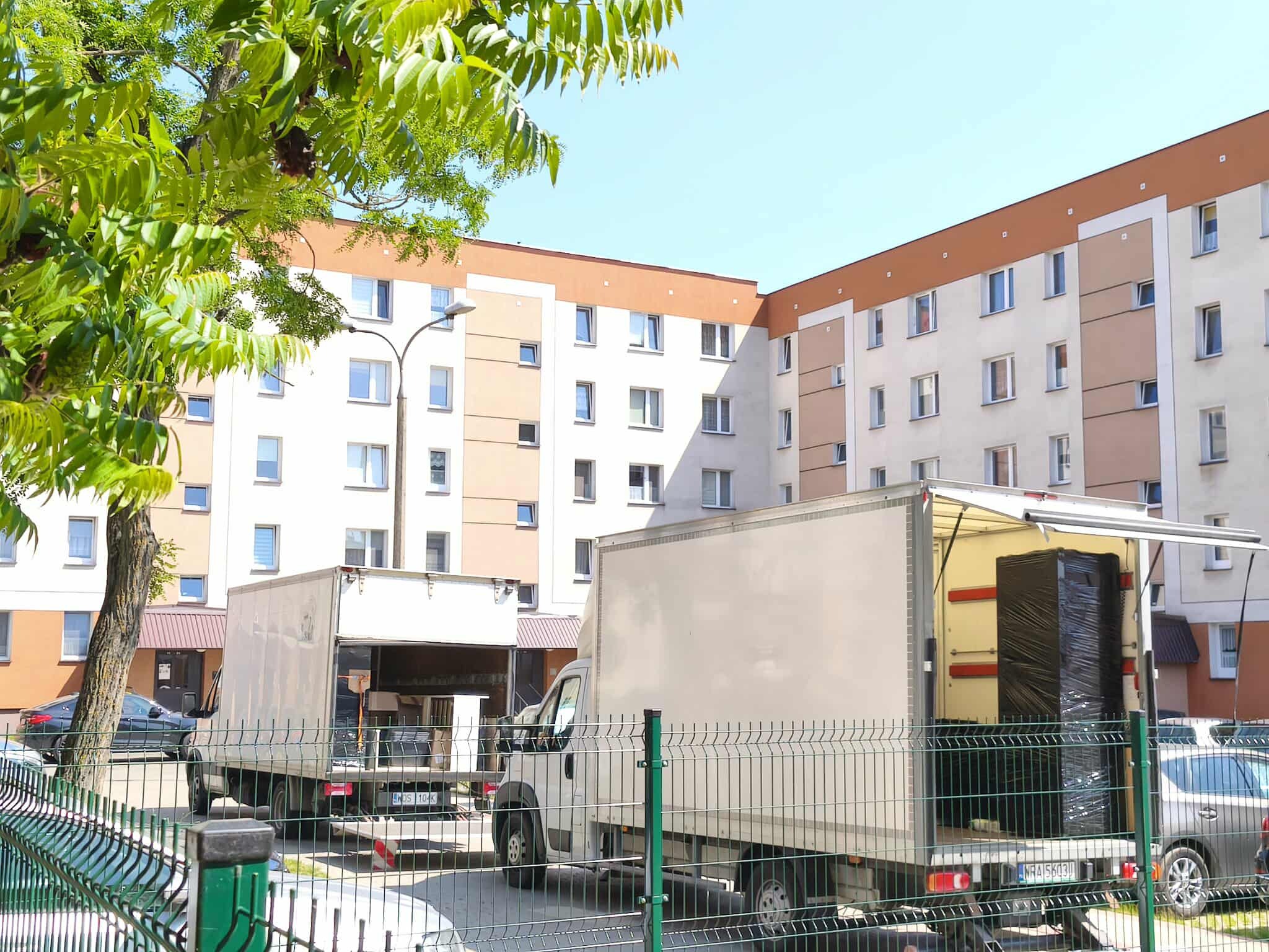 Dwa busy transportowe z otwartymi przestrzeniami ładunkowymi, zaparkowane przed blokiem mieszkalnym w słoneczny dzień. Jeden bus załadowany meblami.