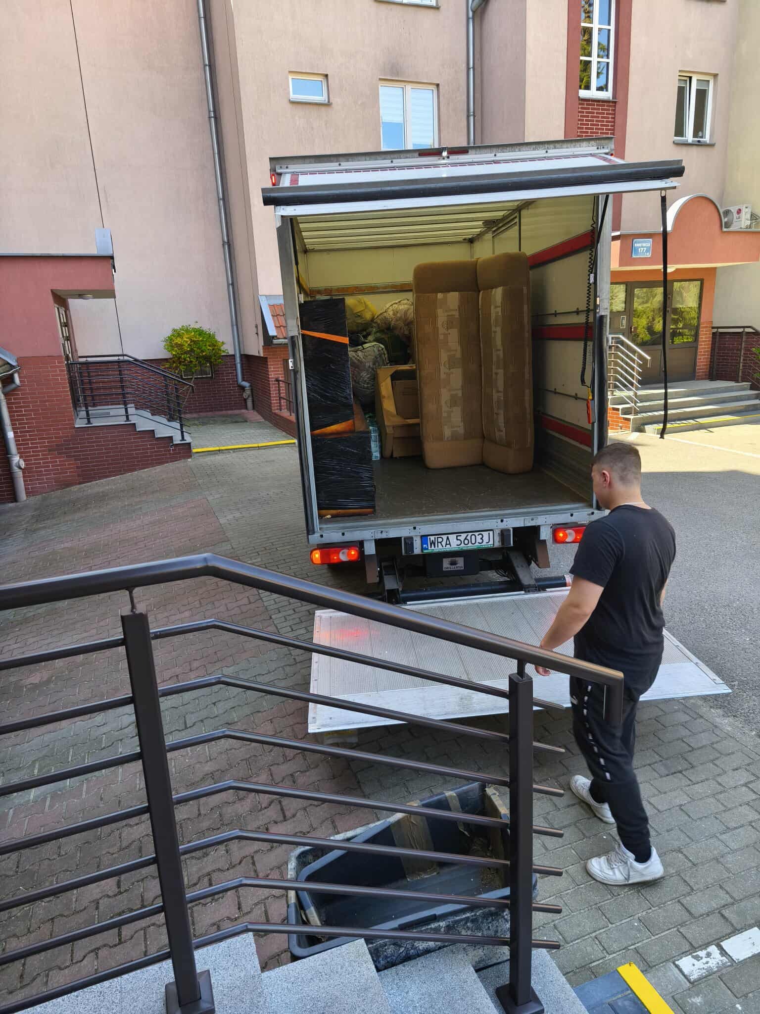 Otwarta paka busa z meblami podczas przeprowadzki. Mężczyzna w czarnej koszulce stoi obok rampy. W tle budynek mieszkalny z numerem 177.