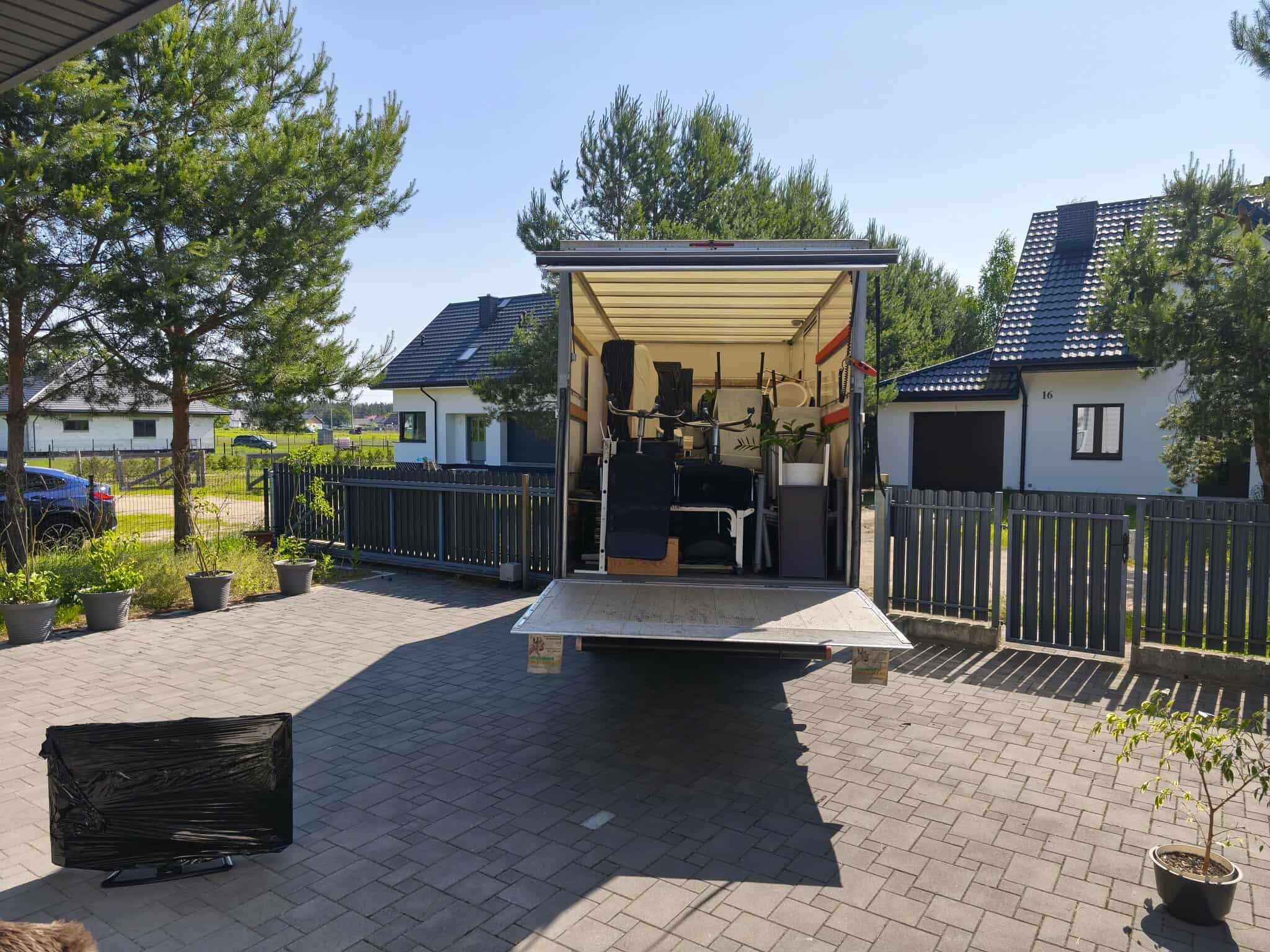 Otwarta paka busa wypełniona meblami i sprzętami podczas przeprowadzki. Na pierwszym planie zapakowany telewizor, w tle domy jednorodzinne i błękitne niebo.