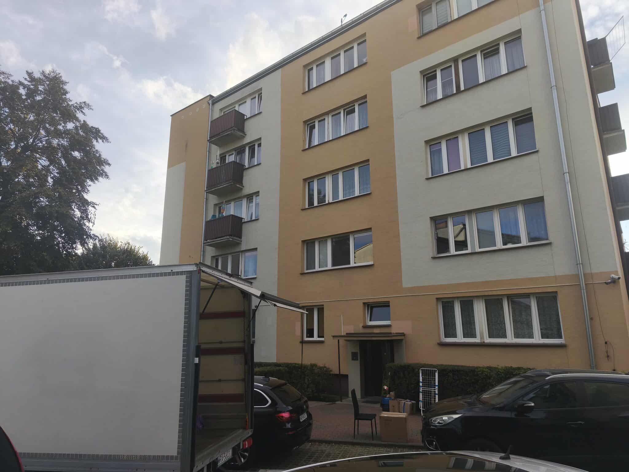 Przeprowadzka: biała ciężarówka z otwartą burtą przed blokiem. Kartony i krzesło na chodniku, zaparkowane auta. Dzień, pochmurne niebo.