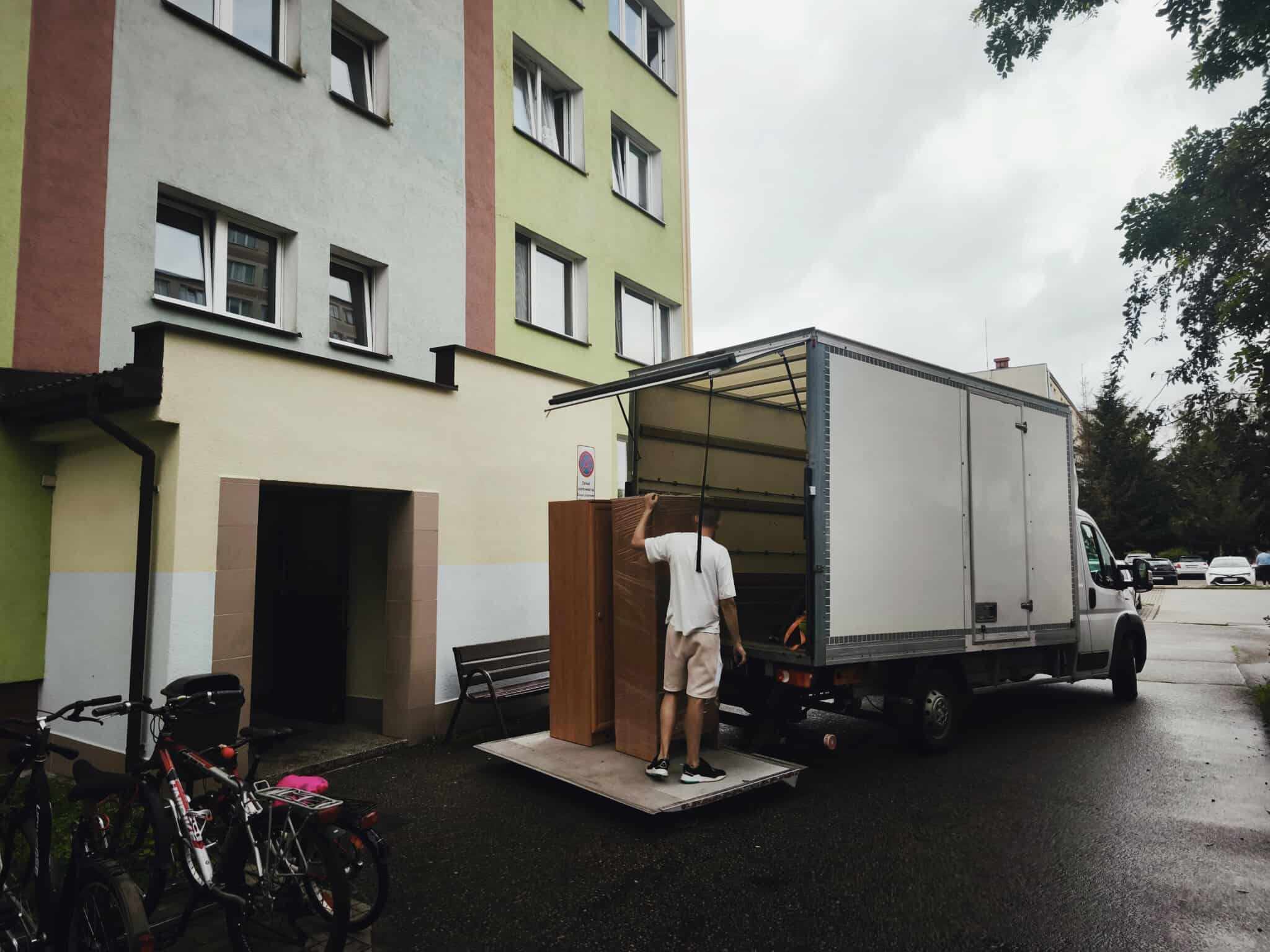 Mężczyzna w krótkich spodenkach wyładowuje owiniętą szafę z białej ciężarówki na tle bloku mieszkalnego. Obok zaparkowane rowery i ławka.