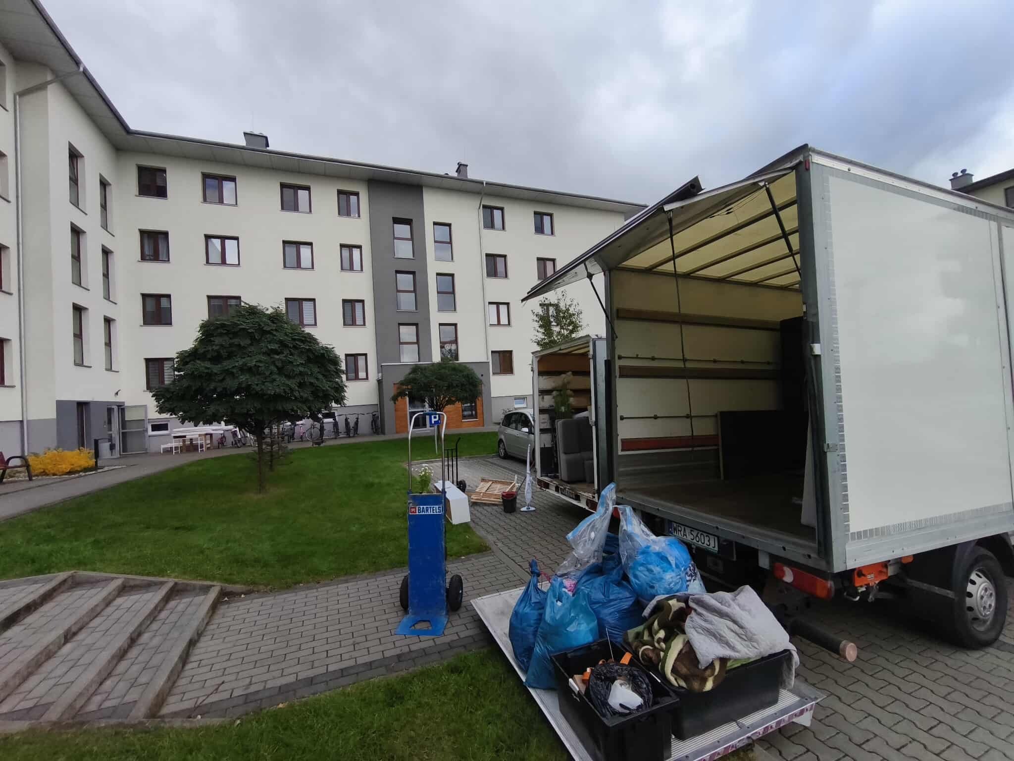 Otwarta paka busa z workami i kartonami na rampie, w tle blok mieszkalny i wózek transportowy. Usługa transportowa w trakcie załadunku.