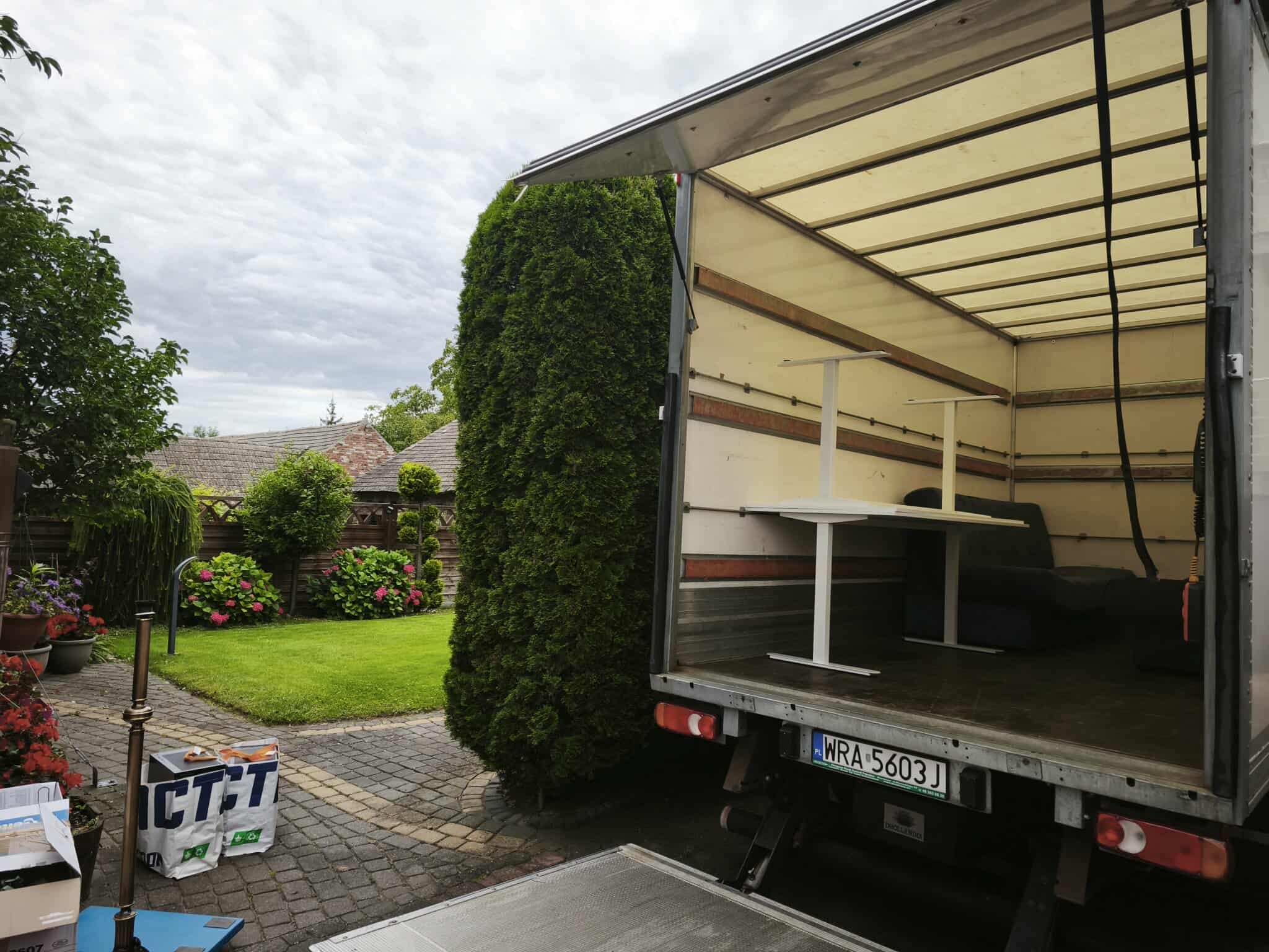 Otwarta paka busa z meblami (stół, sofa) na tle zadbanego ogrodu z tujami i kostką brukową. Widoczne torby z napisem. Tablica rejestracyjna WRA 5603J.