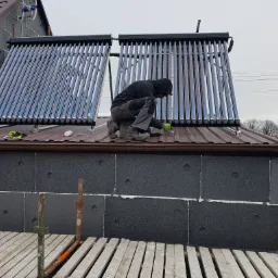 Montaż kolektorów słonecznych na dachu domu z ociepleniem zewnętrznym, widoczne narzędzia i drabina.