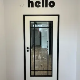 Białe drzwi z czarną ramą i szybą, widok na wnętrze z parkietem. Nad drzwiami czarny napis 'hello' na białej ścianie. Minimalistyczny design.