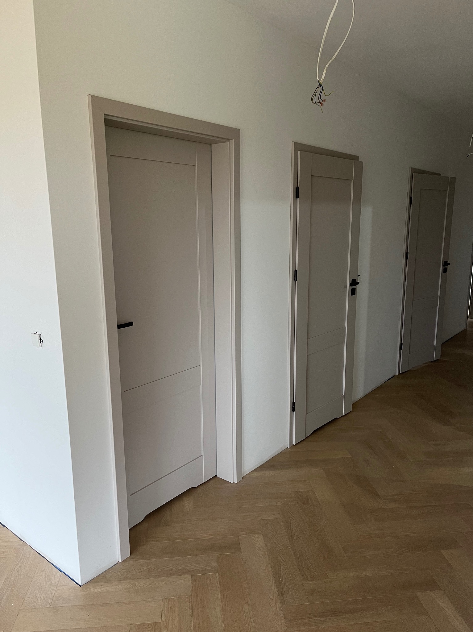 Nowo zamontowane, szare skrzydła drzwiowe z czarnymi klamkami w jasnym wnętrzu z parkietem ułożonym w jodełkę; widoczne wystające przewody elektryczne.