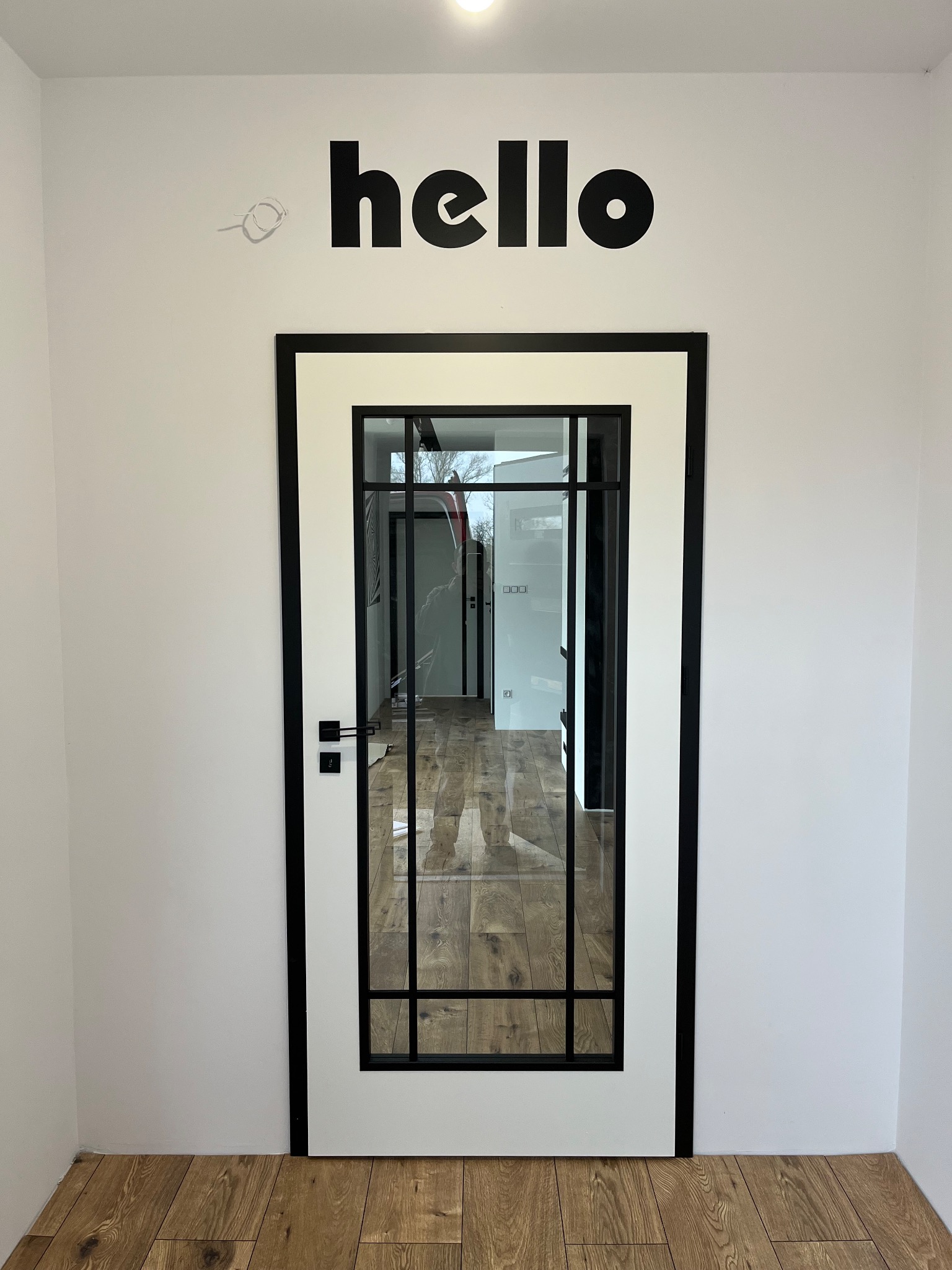 Białe drzwi z czarną ramą i szybą, widok na wnętrze z parkietem. Nad drzwiami czarny napis 'hello' na białej ścianie. Minimalistyczny design.