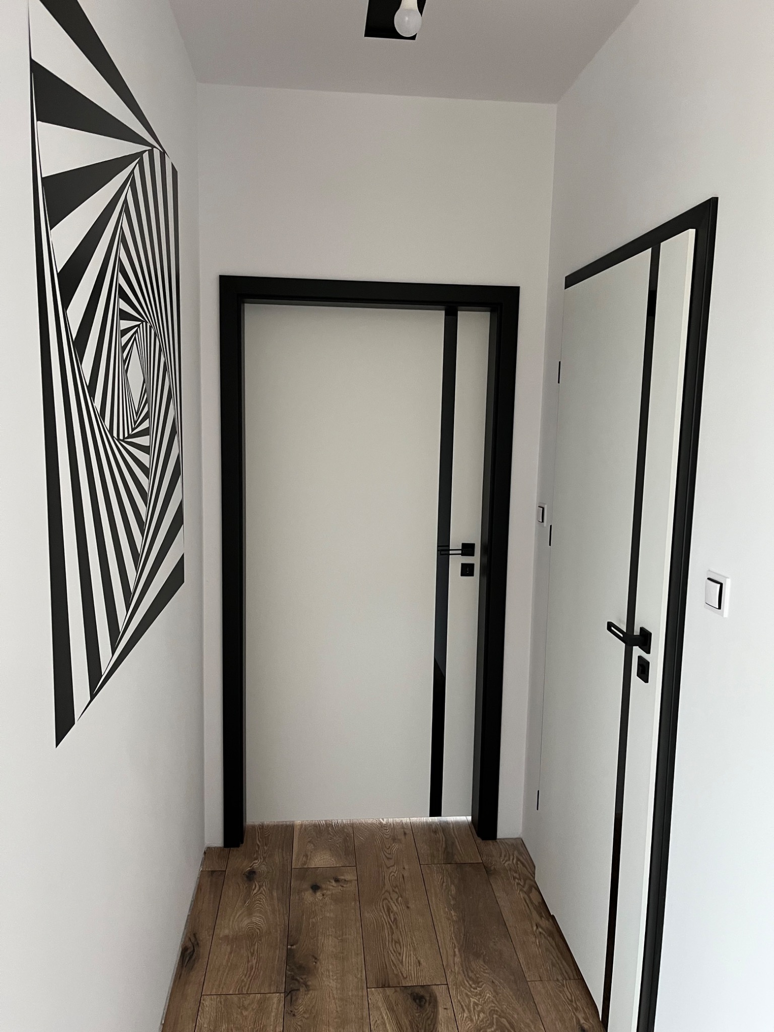 Nowoczesne, białe drzwi wewnętrzne z czarną ościeżnicą w korytarzu z drewnianą podłogą i geometryczną grafiką na ścianie. Minimalistyczny design, eleganckie wykończenie.